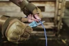 Hands in tactical gloves using a blue multi-tool knife to cut a blue wire outdoors.