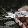 Hand using a stainless steel multi-tool shear to cut a thin wooden branch outdoors.