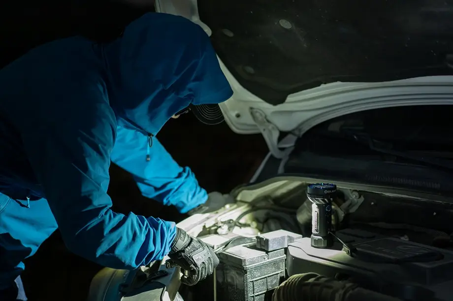 Person in blue jacket and gloves uses Olight flashlight to illuminate car engine repair.