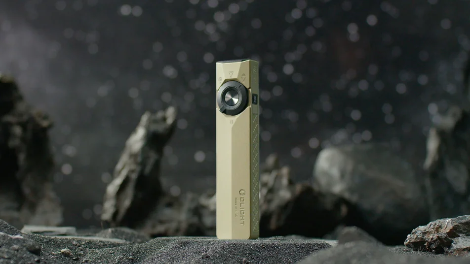 Olight Arkfeld flat EDC flashlight in tan, standing on dark rocky terrain with a blurred starry background.
