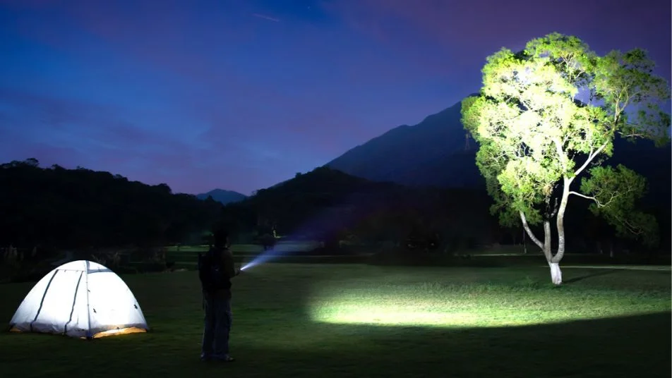 Night camping: person shines flashlight on tree, tent glows. Mountains in background under dark sky.