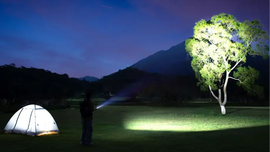 Night camping: person shines flashlight on tree, tent glows. Mountains in background under dark sky.