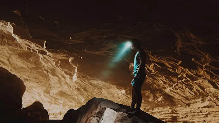 Person with headlamp exploring dark cave, light illuminates textured rock formations.