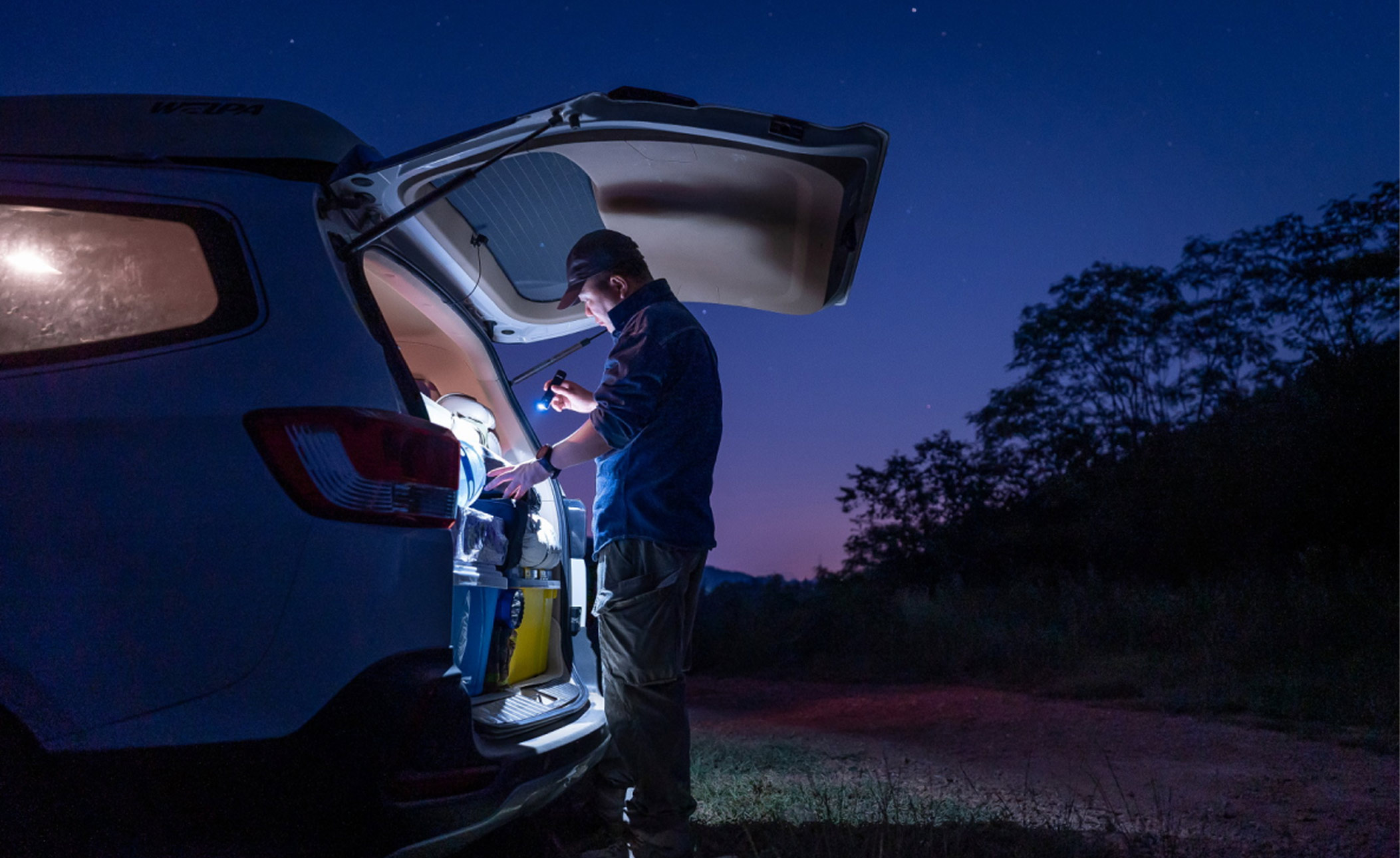 A father outdoors at night with an Olight flashlight from a car trunk. Text: 'Camping, hiking, or just walking the dog.'